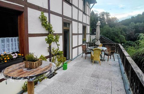 Une terrasse confortable avec une table et des chaises, entourée de plantes. En arrière-plan, un paysage verdoyant est visible.