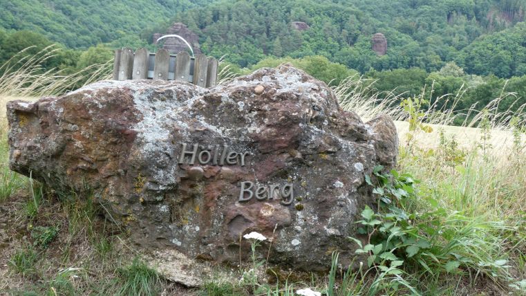 Ein markanter Felsen mit der Inschrift "Holler Berg" steht in einer grünen Landschaft. Im Hintergrund sind sanfte Hügel und Bäume zu sehen.