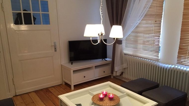 Un espace de vie confortable avec une télévision, une table basse et une lampe moderne. Les fenêtres sont équipées de rideaux et de stores, et le sol est en bois.
