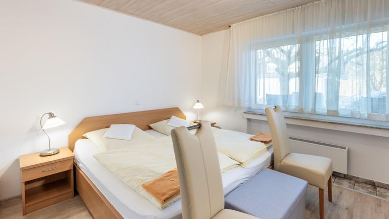 Ein einfaches Schlafzimmer mit einem Doppelbett und hellen Möbeln. Große Fenster sorgen für viel Tageslicht.