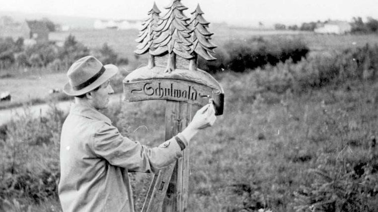 Een man schildert een bord met de tekst "Schlumath". Op de achtergrond zijn groene weilanden en bomen te zien.