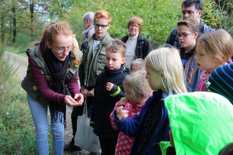 Eine Gruppe von Kindern und Erwachsenen steht im Freien und beobachtet gespannt eine ältere Schülerin, die etwas in der Hand zeigt. Die Szene findet in einer grünen, natürlichen Umgebung statt.