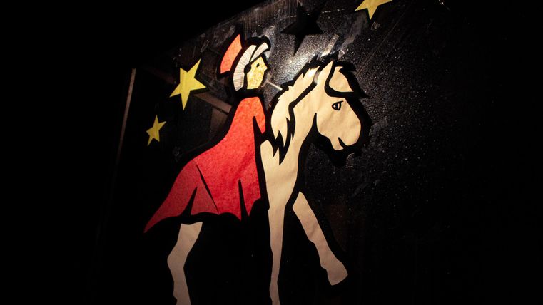 Eine kreative Papierausschnitt-Darstellung einer Figur mit einem Pferd, umgeben von gelben Sternen. Die Figur trägt ein rotes Kleid und einen Hut.