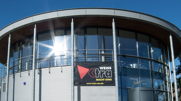 Ein modernes Gebäude mit großen Fenstern und einem geschwungenen Dach. An der Fassade hängt ein Banner mit dem Schriftzug "WEISS xtra macht Kino".