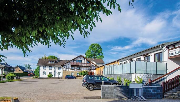 Ein traditionelles Gebäude mit einer Mischung aus Fachwerk und modernem Design. Es liegt an einer Straße und ist von Bäumen umgeben.