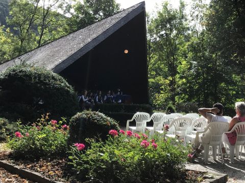 Eine Bühne im Freien, umgeben von Bäumen und Blumen. Im Hintergrund sitzen Menschen in weißen Stühlen.