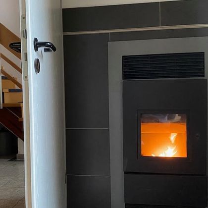 Ein moderner Kaminofen mit flammendem Feuer steht in einem Raum. Eine offene Tür führt zu einer Treppe im Hintergrund.