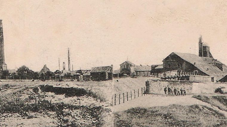 Eine historische Industrieansicht mit Fabrikgebäuden und hohen Schornsteinen. Im Vordergrund ist eine unebene Landschaft mit Zäunen und wenigen Personen zu sehen.