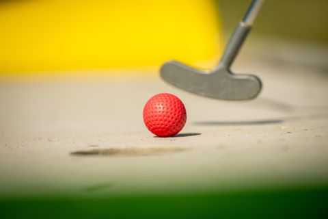 Een rode golfbal ligt op de grond. Een putter is op de voorgrond zichtbaar.