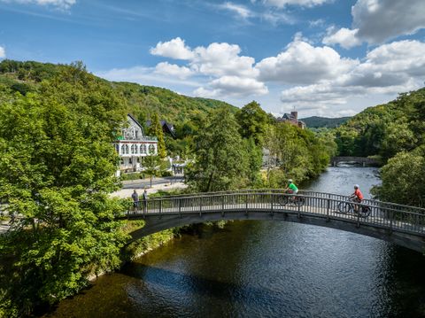 Zwei Radfahrende überqueren eine Brücke über die Rur, umgeben von grüner Landschaft und historischen Gebäuden. Im Hintergrund ist eine Burg auf einem Hügel zu sehen.