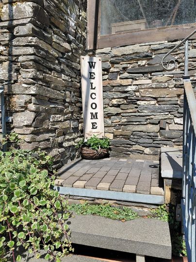 Une entrée de maison accueillante en pierre avec un panneau de bienvenue. Des pots de fleurs et des plantes vertes créent une atmosphère amicale.