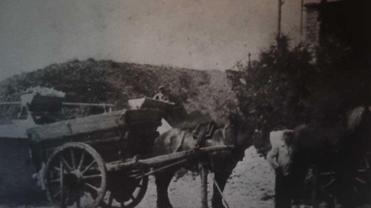 Ein Pferdefuhrwerk steht still auf einer Straße. Der Wagen ist mit verschiedenen Gütern beladen und von Pferden gezogen.