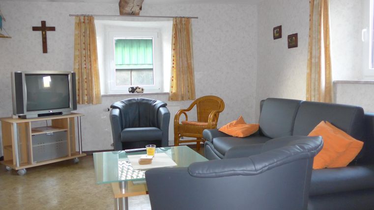 Ein gemütliches Wohnzimmer mit einem schwarzen Sofa und einem blauen Sessel. Den Hintergrund schmücken Vorhänge und ein Fernseher.