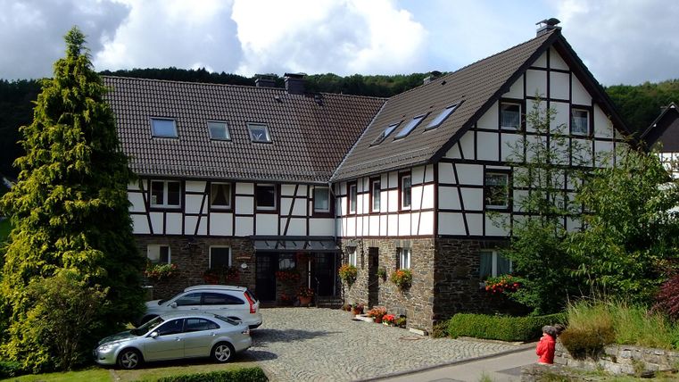 Ein charmantes Gebäude im Fachwerkstil mit einem großen Garten. Vor dem Haus stehen ein paar Autos und es gibt grüne Bäume im Hintergrund.