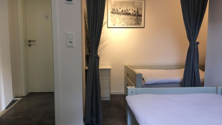 Een eenvoudige kamer met twee bedden en een raam. Gordijnen scheiden de ruimte, en aan de muur hangt een zwart-wit foto.