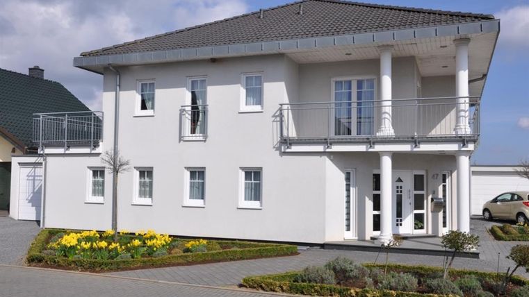 Ein modernes, zweigeschossiges Haus mit einem grauen Dach und großen Fenstern. Der Vorgarten ist mit gelben Blumen und einem schmalen Weg gestaltet.