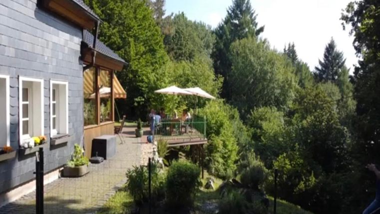 Een gezellig huis omringd door bomen en groen. Op de achtergrond is een terras met parasols en stoelen te zien.