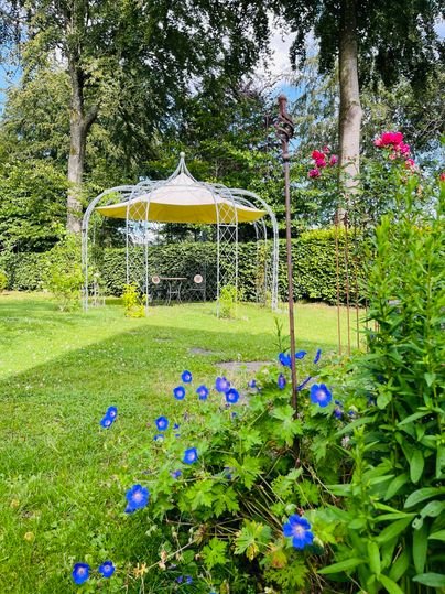 Ein schöner Garten mit bunten Blumen und einem Pavillon im Hintergrund. Die Sonne scheint durch die Bäume und schafft eine einladende Atmosphäre.