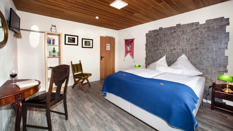 Ein modernes Hotelzimmer mit einem großen Bett und blauer Bettwäsche. Es gibt einen Tisch, einen Fernseher und eine angenehme Holzdecke.