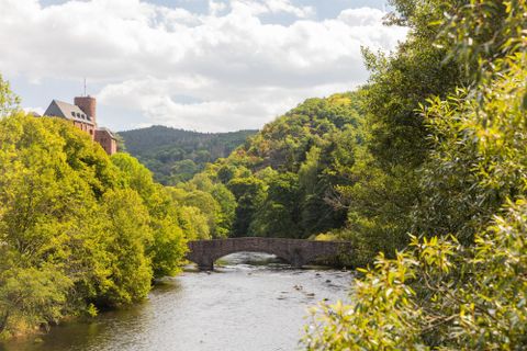 Eine ruhige Flusslandschaft mit einer Steinbrücke und umgeben von üppigem Grün. Im Hintergrund sind sanfte Hügel und ein Gebäude sichtbar.