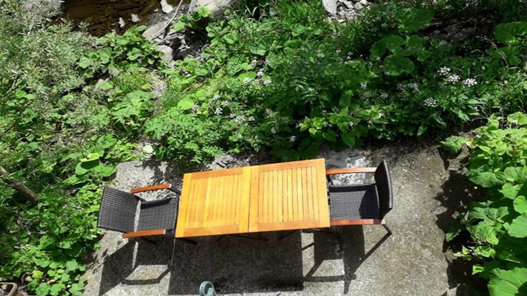 Une belle table avec deux chaises se trouve au milieu des plantes vertes. En arrière-plan, un petit ruisseau coule.