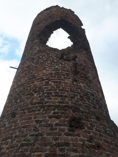 Ein alter, zerfallener Turm aus roten Ziegeln mit einem großen Loch an der Spitze. Der Himmel ist bewölkt und es wirkt trostlos.