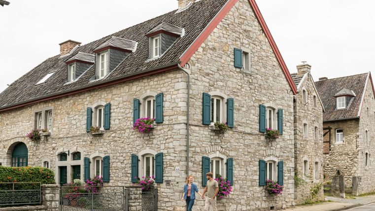 Ein charmantes Steinhaus mit Fensterläden in Blau und blühenden Pflanzen. Zwei Personen stehen auf dem Gehweg und unterhalten sich.