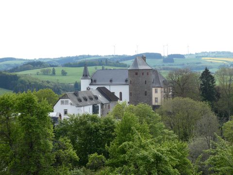 Un vieux château entouré d'un paysage verdoyant et d'arbres. À l'arrière-plan, on peut voir des collines douces et des éoliennes.