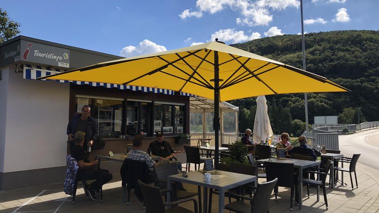 Ein gemütliches Café im Freien mit einem großen gelben Sonnenschirm. Gäste sitzen an Tischen und genießen die Sonne.