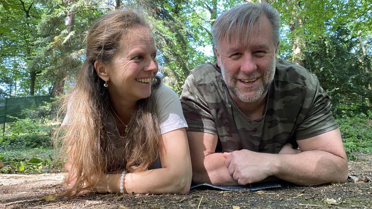 Ein Paar liegt lächelnd auf dem Boden im Wald. Um sie herum sind grüne Bäume und eine sonnige Atmosphäre.