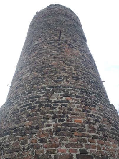 Ein historischer Steinbau, der hoch in den Himmel ragt. Der Turm hat eine rustikale Struktur aus Ziegelsteinen und ist von einer grauen Wolkenlandschaft umgeben.