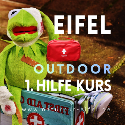 Ein Puppen-Greenie mit einem Erste-Hilfe-Kasten sitzt auf einer Kiste. Der Text deutet auf einen Outdoor Erste-Hilfe-Kurs in der Eifel hin.