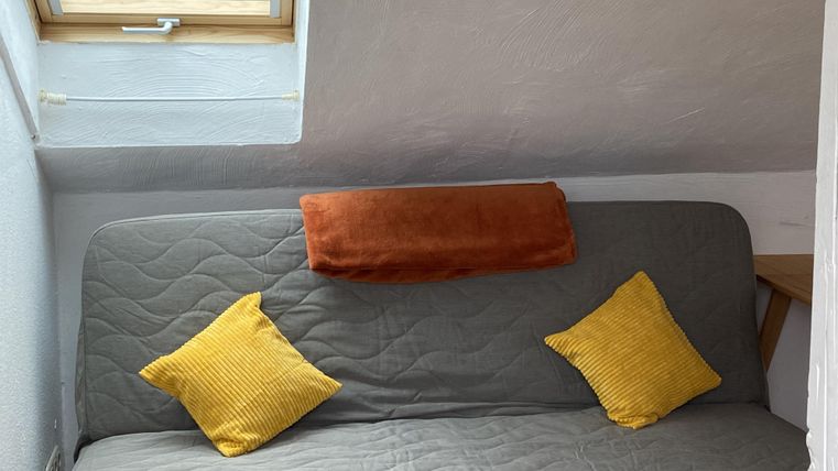 Een comfortabele slaap bank met een grijze bekleding en drie kussens in verschillende kleuren. Een raam zorgt voor daglicht in de kamer.