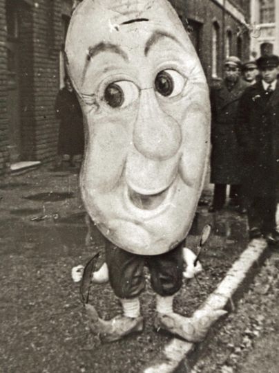 A person wearing a large costume mask is smiling. In the background, several people are standing in historical clothing.