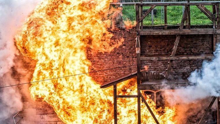 Ein intensives Feuer bricht aus und umgibt ein Holzgebäude. Dichter Rauch steigt auf und erzeugt eine dramatische Atmosphäre.