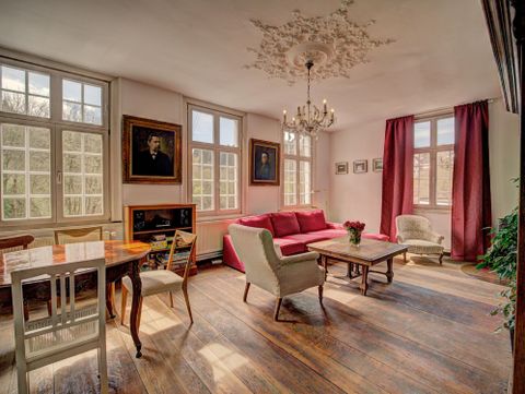 A cozy living room with large windows and a beautiful wooden floor. There is a red sofa, a dining table, and decorative pictures on the walls.