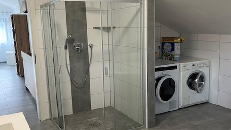 Ein modernes Badezimmer mit einer Glastür-Dusche und einer Waschmaschine neben einem Wäschetrockner. Der Boden ist mit grauen Fliesen ausgelegt und die Wände sind hell gefliest.