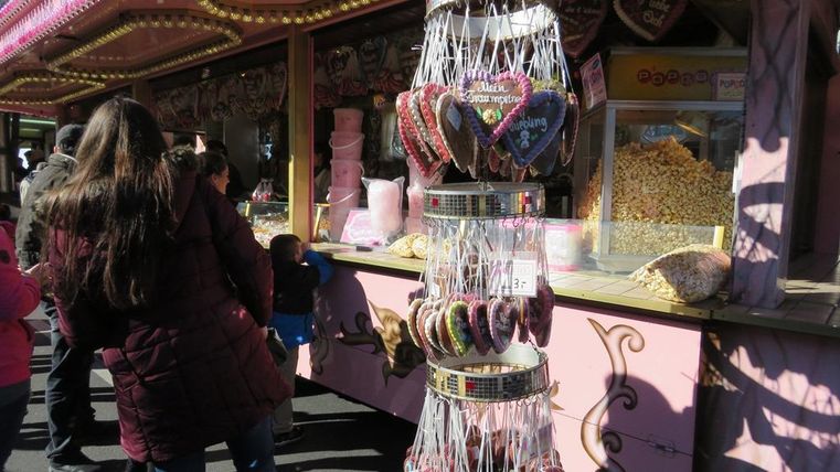 Ein bunter Verkaufsstand mit Zuckerherzen und Süßigkeiten. Menschen stehen in der Nähe und schauen sich die Leckereien an.
