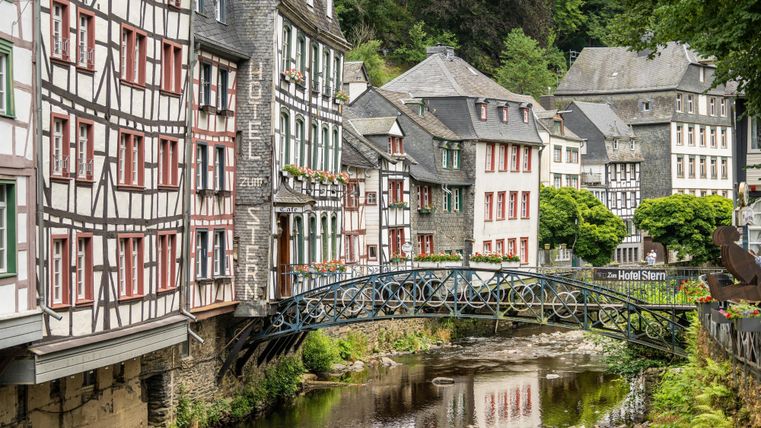 Eine malerische Stadtansicht mit Fachwerkhäusern und einer kleinen Brücke über einen ruhigen Fluss. Üppiges Grün umgibt die historische Architektur.