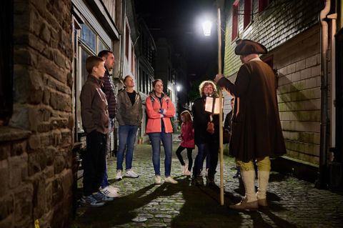 Eine Gruppe von Menschen hört einem Geschichtenerzähler in historischer Kleidung in einer schmalen, beleuchteten Gasse zu. Es ist Nacht und die Atmosphäre ist freundlich und einladend.