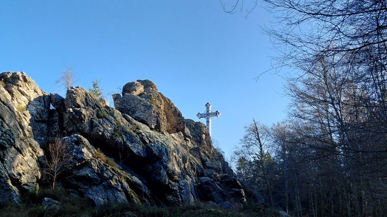 Ein schöner Felsen mit einem Kreuz, umgeben von Bäumen und klarem Himmel. Der Pfad führt durch die Natur.