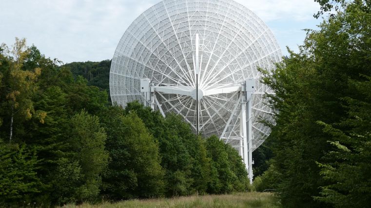 Eine große Satellitenschüssel steht zwischen Bäumen in einer grünen Landschaft. Der Himmel ist teilweise bewölkt.