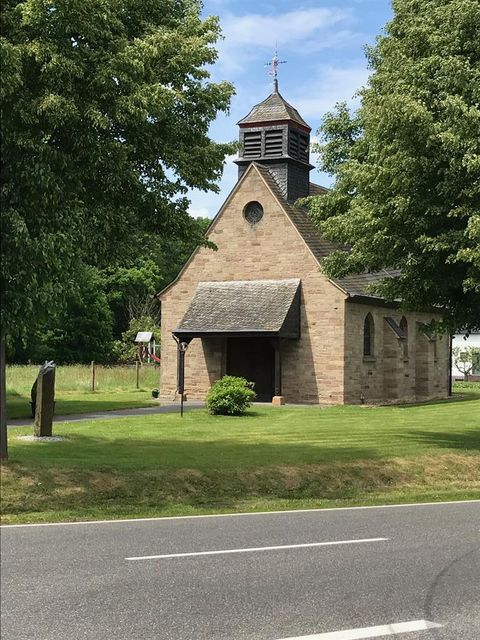 Eine kleine Kirche aus Stein umgeben von Bäumen und grünem Gras. Der Himmel ist klar und blau.