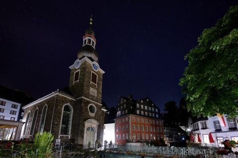 Eine historische Kirche mit einem markanten Turm steht bei Nacht im Vordergrund. Im Hintergrund sind weitere alte Gebäude und Bäume zu sehen.
