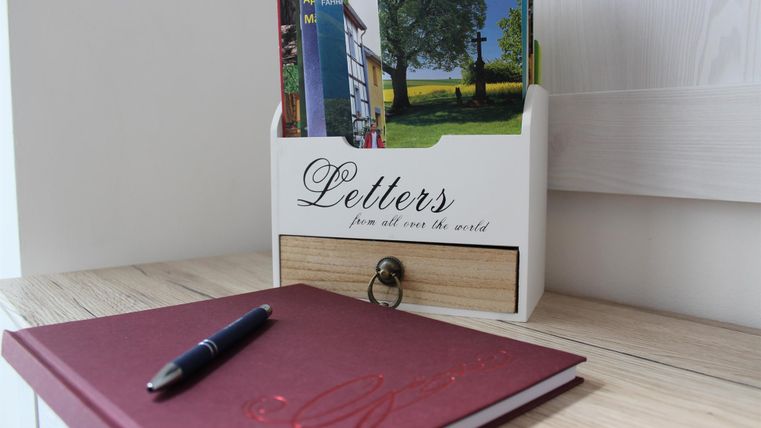 Ein Holzregal mit Postkarten und einem Notizbuch auf einem Tisch. Daneben liegt ein Kugelschreiber.