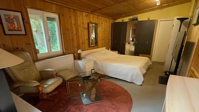 Een gezellig kamer met houten wanden en een bed. Er is een fauteuil, een tafel en grote ramen die licht binnenlaten.