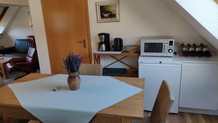 Ein gemütlicher Essbereich mit einem Tisch, Stühlen und einer Vase voller Lavendel. Im Hintergrund sind eine Mikrowelle, eine Kaffeemaschine und Regale mit Weinflaschen zu sehen.