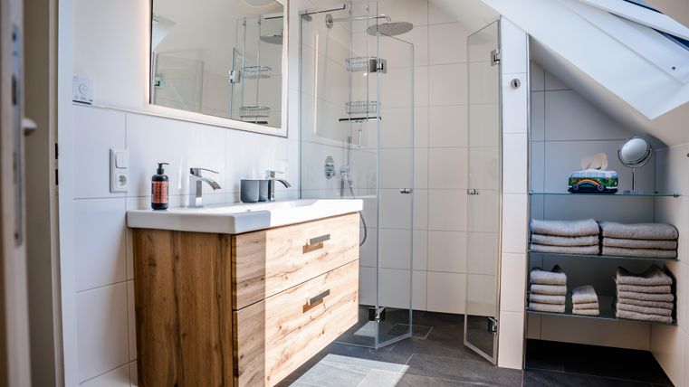 Ein modernes Badezimmer mit einer Dusche aus Glas und einem Holzwaschtisch. Handtücher sind ordentlich auf einem Regal gestapelt.