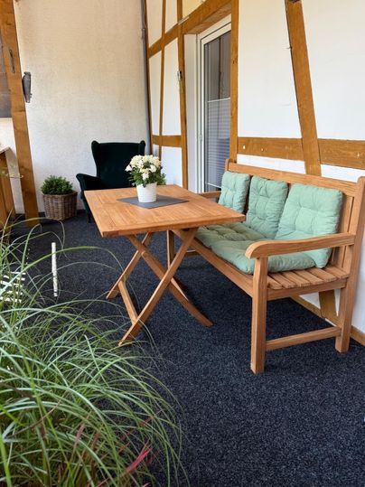 A cozy seating area on a balcony with a wooden table and a bench. On the table, there is a small flower vase, and in the background, plants are visible.