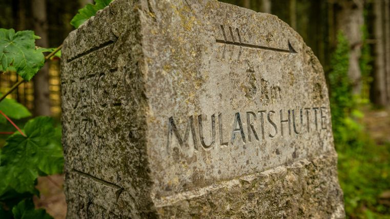 Een stenen wegwijzer met het opschrift "MULARTHÜTTE". Omringd door bomen en plantaardig groen.